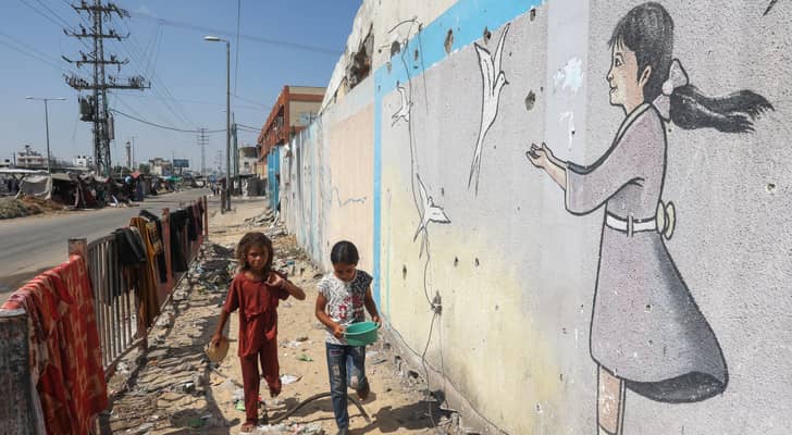 فتيات فلسطينيات يسيرن أمام جدارية في مخيم نازحين مؤقت أقيم على جانب الطريق في دير البلح وسط قطاع غزة